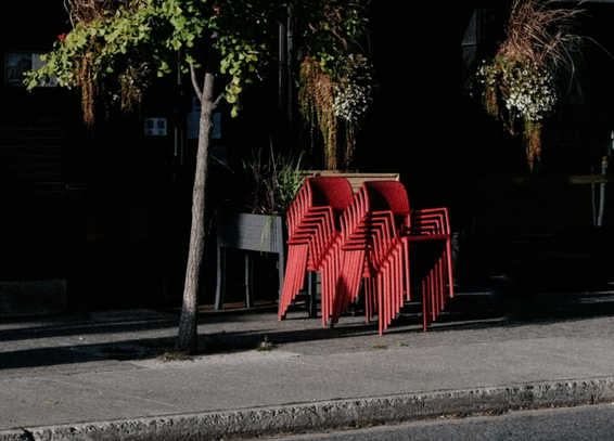 Two stacks of bright red plastic chairs are seen on the sidewalk outside a dark building. The early evening light highlights the chairs as well as a small tree and two hanging planters.