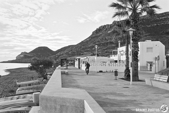 A peaceful coastal promenade with palm trees, lined by white buildings and rocky hills in the background. A person rides a bicycle along the path, while another stands nearby, creating a serene and picturesque scene.