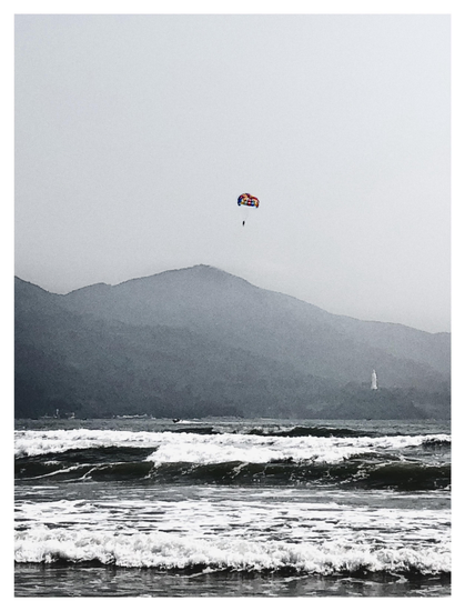 A view of a beach with large waves crashing ashore in the foreground. In the background are low mountains, at the foot of which a white marble statue of a woman stands, enormous in reality but made tiny by the distance. A parasail bearing a lone figure hangs in the sky above. The image has been desaturated except for the vivid rainbow colors of the parasail chute, which pop against the monochrome landscape and featureless sky.