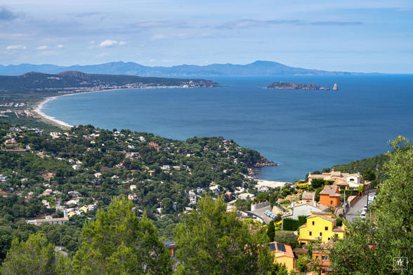 Color photo taken from a high point on a sunny day looking up a curving coastline on the Mediterranean Sea with colorful houses in the foreground, some small islands offshore in the middle distance, and low mountains on the horizon. 