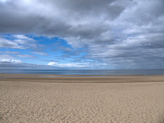 A minimalist landscape of a beach on sunny day. The foreground is dominated by a vast expanse of beige sand that stretches toward a calm blue sea in the distance. The top half of the image is taken up by a wide, cloudy blue sky.