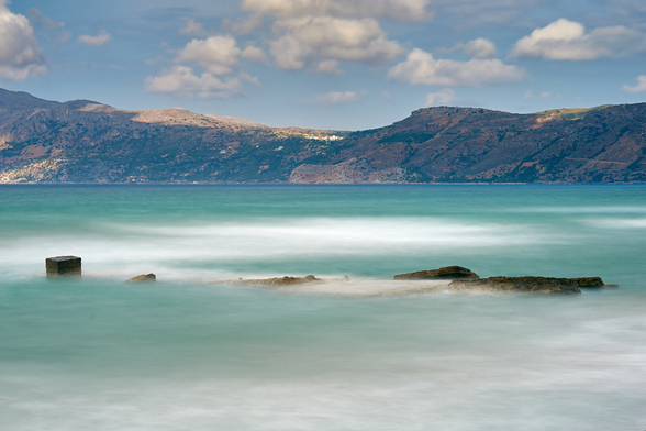 The photo shows a long-exposure of the sea. The sea looks very milky, mostly turquoise greenish in the front with white patches and blue in the back. In the front some cement fragments are seen. In the background the eastern end of the gulf in form of mountains are visible. Above the mountains is the blue sky with white clouds.