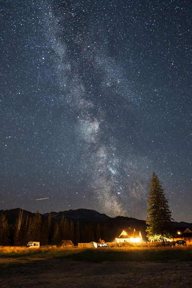 Night long-exposure shot of the Milky Way stretching vertically across a star-filled sky above the Tatra Mountains near Kościelisko, Zakopane, Poland, with a warmly lit wooden highland cottage, conifer trees, and dark mountain silhouettes in the foreground.