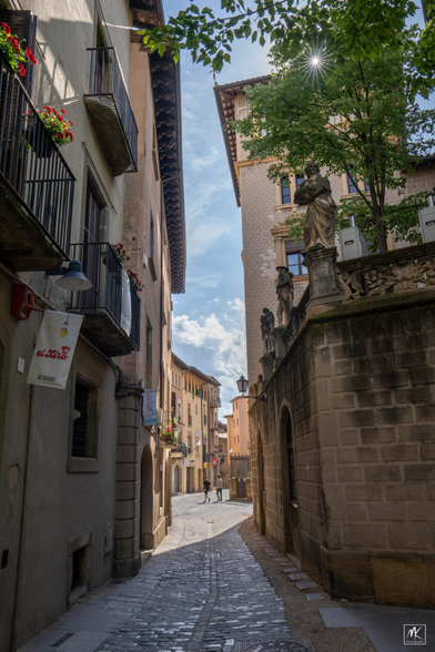 Color photo looking down a narrow cobblestone street in a Catalonian city center that is shaded in the foreground and sunlit beyond. To the right is a wall with statues of people on top of it and above them the is a rayed sunburst peeking through tree branches. In the distance are two people walking down the street.  