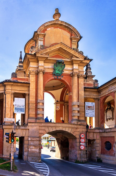 This is a photo of the Arch of Meloncello. It is an ornate Baroque archway built over a road street. This monumental structure has warm terracotta and ochre colours, columns, walkways on an elevated level (the stairs up to San Luca), and a green coat of arms centered above the main upper arch. Below the pedestrian section there is a stone arch which makes a tunnel that allows a paved road with traffic signs to pass underneath. A few people are walking through the elevated portion.