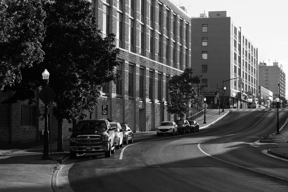 A road in a city is seen curving to the right and heading up a slight hill towards traffic lights. Large buildings are seen along the left side of the street and some parked vehicles and small trees are also seen. This is a black and white image.