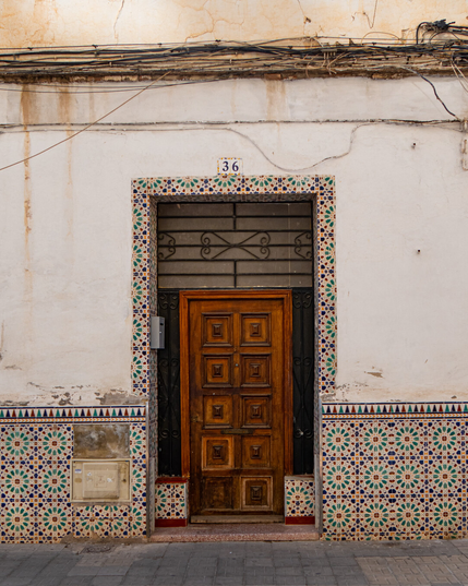 A symmetrical photo of an entranceway in Málaga, Spain. A dark, paneled wooden door sits in a shaded recess, framed by a weathered white plaster wall with colorful, geometric ceramic tiles running along the base. Overhead, a tangle of cables stretches across the facade, just above a ceramic house number tile reading '36'.
