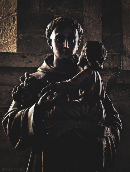 A dark wooden statue depicting two figures set against a textured stone wall background. 

A man dressed in traditional monastic robes with a prominent cowl or hood resting around his shoulders. He has short hair with a tonsure. He is looking forward with a solemn expression.

The man is holding a young child in his arms. The child is depicted in profile, looking up toward the man’s face.