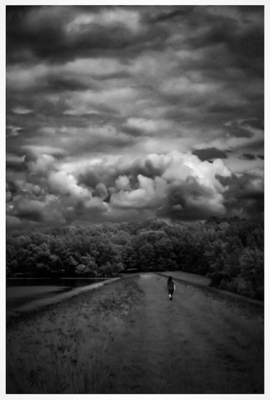 Black and white landscape photo of a path on a levee going away from the camera. Someone in the distance is walking away on that path towards a tree line. Above the tree line dramatic clouds can be seen. Some of the clouds kind of look like a giant eye.