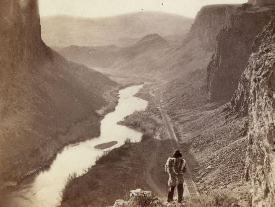 d'un point escarpé, un amérindien regarde la voie ferrée longeant la rivière entre les montagnes sauvages
