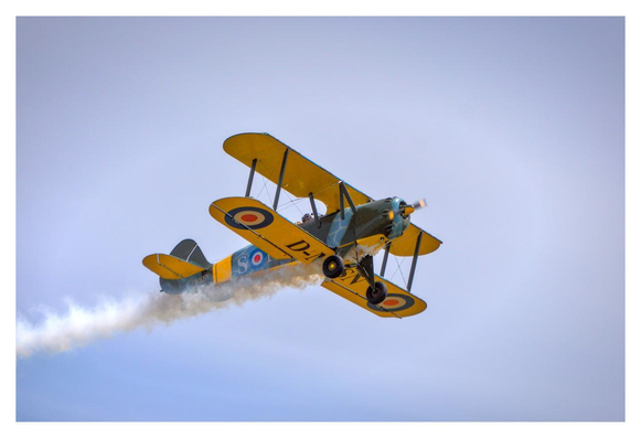 A vintage yellow biplane is flying at a steep upward angle against a clear, bright blue sky. The aircraft is a classic open-cockpit double-winged (biplane) design with bright yellow wings, while the the tail and front fuselage has a dark green/blue camouflage pattern. British Royal Air Force roundels (concentric circles in red, white, and blue) are clearly visible on the wings and fuselage. The markings "D-A" can be seen on the side of the aircraft. A long, thick trail of white smoke streams behind it, curving downward from the tail. The pilot is visible sitting in the open cockpit. The entire scene is captured from a low angle, emphasizing the dynamic upward movement of the aircraft.