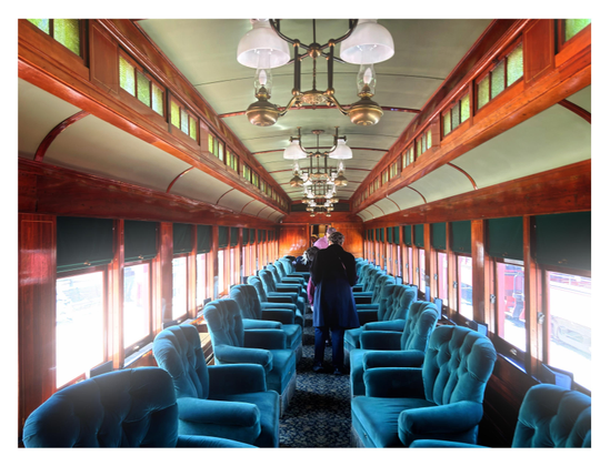 Photo looking down the carpeted center aisle of a vintage train dining car. To the left and right are rows of windows with raised green shades, letting in a glare of sunlight. In front of the windows are comfortable-looking upholstered blue armchairs facing inward towards the aisle but capable of swiveling 360 degrees. Small menus are set up on the window ledges beside each chair. Above is a row of chandeliers in gold casings suspended from the green ceiling, flanked by two upper rows of smaller windows with greenish panes enclosed by wide borders of warm wood. All the lines in the photo recede to a central vanishing point. In the middle of the aisle are a couple of standing passengers, getting settled in.

This train is part of the historic Strasburg Rail Road, which continues to offer short rides along a 9-mile route with station stops throughout a stretch of Amish country in Lancaster County, PA.