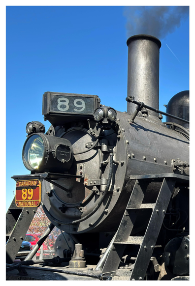 Closeup of the front of a sunlit steam locomotive, with its black main cylinder studded with rivets, round headlight, and chimney belching black smoke. A metal sign above the headlight reads “89”; a smaller red plate underneath the light also reads “89” surrounded by the words “Canadian National.” Through some of the engine’s parts we can glimpse parked cars and trees. The sky above is a vivid, clear blue.