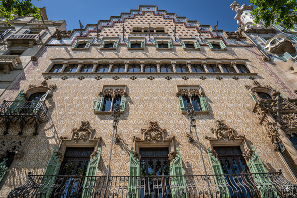 Color photo looking up the facade of a building done in the Moderisnme style with carved patterns on the terracotta-colored wall between ornate widows with green shutters.  At the top are more rows of windows beneath a stepped gable.