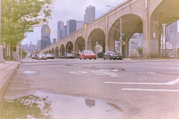 A puddle reflects the street scene by a main road. The Manhattan skyline is in the distance. The road is next to an elevated rail line for the 7 train, a low concrete viaduct with rounded arches.