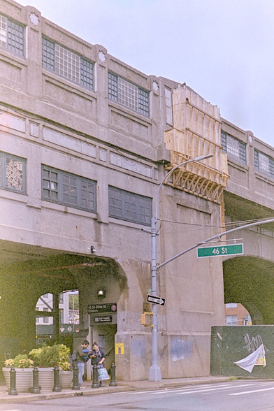 46th St 7 train station, showing staircase leading up to the elevated station on a concrete viaduct. Two people were conversing by the entrance.