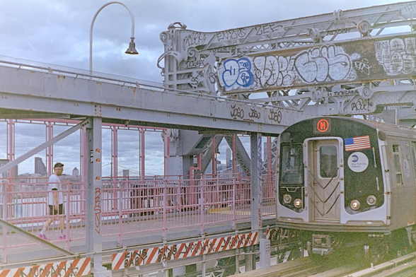 M train beneath steel beams full of graffiti, in front of the pedestrian side of the bridge