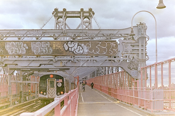 M train coming towards me on the track next to a bike path, beneath giant steel beams covered in graffiti