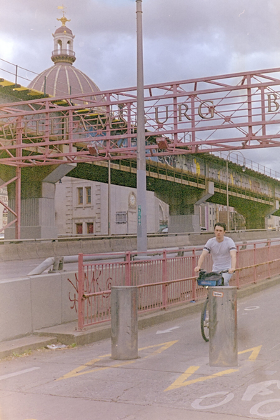 A man riding an analog CitiBike descending from a bridge. In the background is a ramp for trains full of graffiti in front of a church dome
