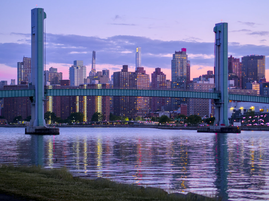Small teal lift bridge in front of many high-rise residential buildings at dusk. The bridge and the buildings are reflected in the river.