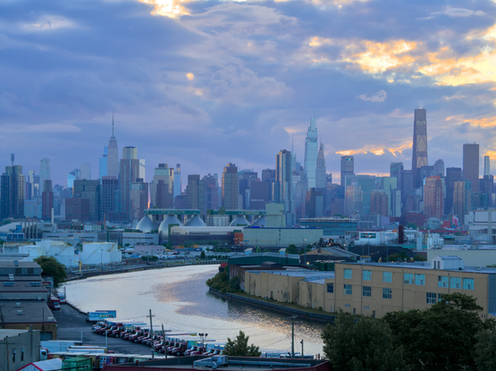 A river curves through an industrial area. The Midtown Manhattan skyline is in the background. Mostly cloudy at dusk.