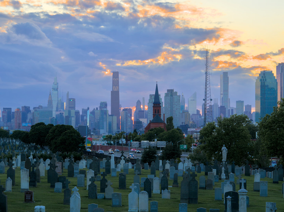 A church built with red bricks, with a tall steeple, above a cemetery. The Midtown Manhattan skyline is in the background. Mostly cloudy, at dusk.