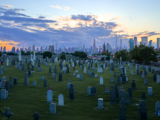 Midtown Manhattan skyline behind a vast cemetery, with many old headstones over green grass. It was partially cloudy, at dusk.