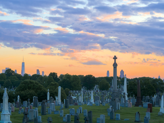 The WTC is in the distance, with cemetery in the foreground. A thin headstone with a cross at the top reaches into the sky, as if rivaling the WTC.