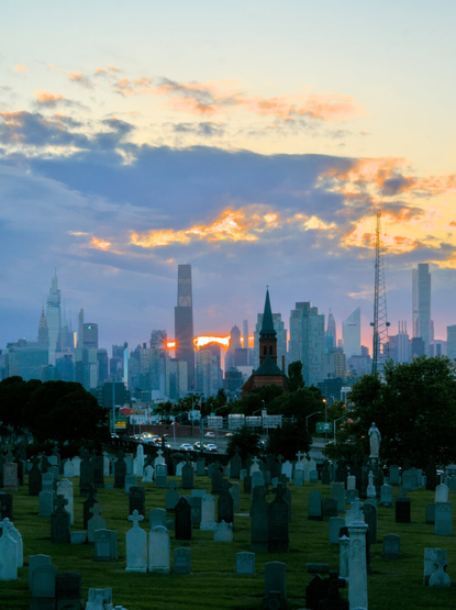 The setting sun peeks out between two skyscrapers, with a cemetery in the foreground.