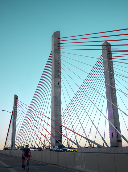 Suspension bridge with modern minimalist concrete towers and red cables; a cyclist was riding below towards me. To the right is a freeway.
