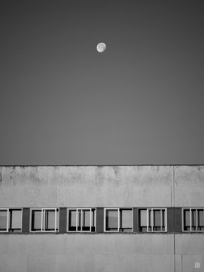 La imagen en blanco y negro muestra la parte superior de un edificio de fachada lisa con varias ventanas alineadas en una sola fila. Sobre el edificio, en el cielo despejado, se ve la luna en una fase próxima a llena. La composición transmite una sensación de simplicidad y calma.