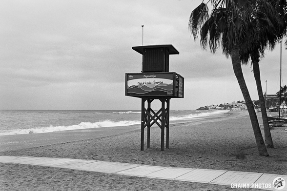 A lifeguard tower stands on a sandy beach beside gentle waves, framed by palm trees under a cloudy sky. The area appears tranquil, ideal for relaxation or beach activities.