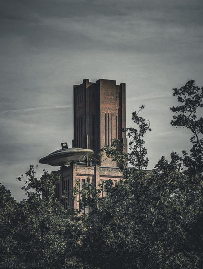A surreal architectural scene featuring a historic brick building (De Inktpot) with an unexpected sci-fi twist.

In the center stands a massive, industrial-style brick building featuring a tall, imposing central tower with narrow, vertical windows. The lower half of the building is heavily framed by thick, dark green tree foliage.

Perched precariously on the roof of the lower section of the building is a large, metallic, silver flying saucer (UFO). It looks as though it has crashed into or landed on the edge of the structure.

The UFO sculpture, titled Zover ("So Far"), was created by artist Marc Ruygrok for an art exhibition in Utrecht and was left on the building permanently.