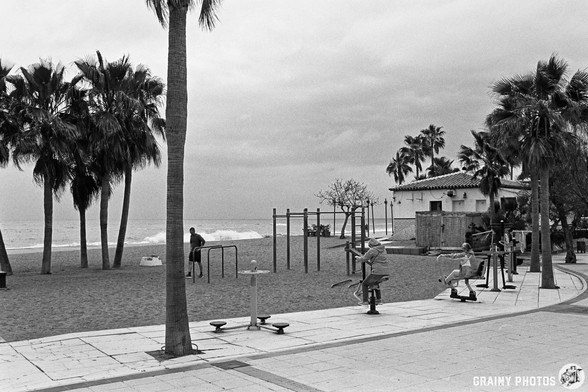 A cloudy beach scene featuring a sandy area lined with palm trees. Two individuals are using outdoor exercise equipment, while another person exercises in the background. A beach restaurant (Chiringuito) is visible near the shoreline.