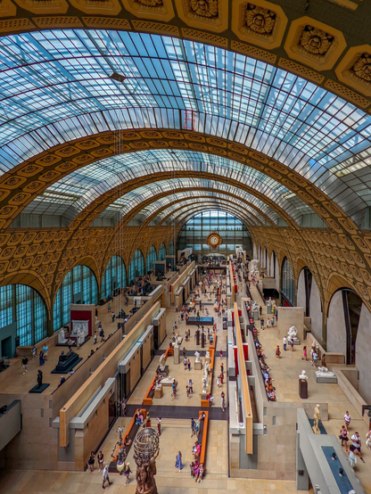 A high, wide-angle shot looking down at the main hall of the Musée d'Orsay. A massive curved glass and iron roof hangs over the interior propped up by towering stone arches with decorative square reliefs. On the floor below, visitors walk around sculptures and displays arranged along a central stone path. A large clock is visible at the far end of the hall.