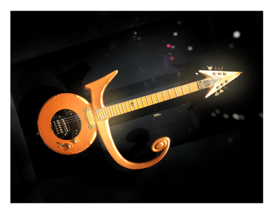 Prince’s “Love Symbol” guitar, with a gleaming gold finish, displayed against a black backdrop behind glass that reflects blurred points of light like stars. The guitar’s shape evokes the wordless symbol that Prince adopted as his name for part of his career: a circular body topped with a lopsided crossbar that extends and swirls upward to the right, and an arrow-shaped headstock with a pointed tip. The guitar stands upright but the image is rotated about 45 degrees which gives it a sense of motion, like a rocket blasting through space.

Displayed at the 2019 exhibition “Play It Loud: Instruments of Rock & Roll” at the Metropolitan Museum of Art in NYC. Details from the museum placard:

"LOVE SYMBOL"
Electric Guitar
Jerry Auerswald, Konstanz, Germany
1993
Maple body, neck, and fingerboard; 25½ in. scale; gold finish on body and neck; EMG single-coil and humbucking pickups, three-way selector switch, volume and tone controls