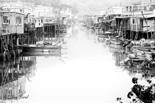This is a period looking photo in landscape format looking along the tidal river of a coastal fishing village with old houses built on wooden stilts on either side of the water. Tai O, Lantau Island, Hong Kong (2014).

The narrow river extends away from the camera into the distance, about one hundred metres away. The river is calm and has a bright silver/white appearance that reflects the surroundings in it's surface. Along either side of the photo are numerous wooden shack-like houses, all built close together and supported by wooden piles driven into the bed of the river. Most of the houses have one floor though one or two have an upper floor. Many of the houses have ladders leading down to small dinghies moored to the wooden piles. The houses, wooden piles and dinghies are reflected on the sides of the river in the water's surface. The centre of the river is overexposed to a featureless white. There is a small dark bush in the lower right corner. After about one hundred metres the river bends to the right and out of view with a lush tropical forrest beyond the bend and with terrain rising out of view, partially obscured by a thick, humid haze. 
