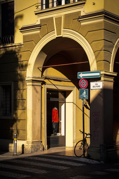 This is a photo of a  street corner featuring a classic Bolognese building with yellow and beige facade stone details and a large archway. Underneath the arch there is a shop window with a mannequin dressed in a bright red jacket and dark pants. On the right hand side there is a pillar with a street sign reading "VIA BELFIORE," along with a blue arrow pointing left. There's also a red and blue no-parking sign. Kinda behind the arch and pillar there is a black bicycle is parked upright against the base of this pillar. In the foreground on the left there is a zebra crossing.