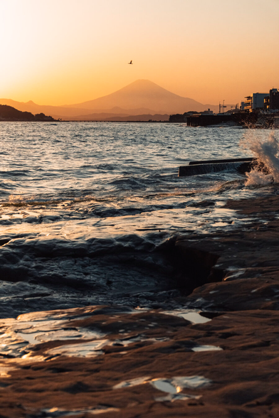 Foreground: wet rocks with water splashing, middle ground: rippling water body, background: Mount Fuji silhouette against warm sunset sky, distant mountains and right-side urban structures near water.