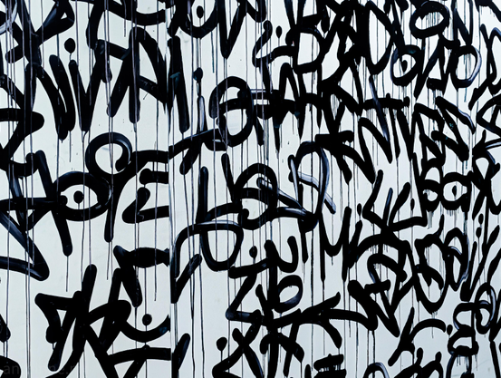 A dense layer of black graffiti tags covering a solid off-white surface. The tags are heavily layered, overlapping each other to create a chaotic, abstract message.

The spray paint is applied heavily, causing numerous distinct vertical drips to run down the length of the wall. These thin, dark streaks add a sense of gravity and raw texture to the piece.