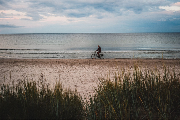 A lone cyclist rides along a flat sandy beach at the water’s edge, a small solitary figure against a wide open Baltic seascape. Tall coastal grass fills the dark foreground, framing the scene from below. The wet sand reflects a pale grey light. The sea is calm and glassy, stretching to a flat horizon under a dramatic sky of layered clouds with breaks of pale blue. The mood is quiet and slightly melancholic. Shot on a Fujifilm X-T30 II with the XF 27mm f/2.8 R WR at f/5.6, 1/160s, ISO 200.