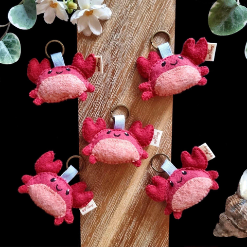 Five little handmade crab keychains photographed on a black and wooden background with a few decorative plants and items. The keychains are attached to bronze key rings with blue cotton ribbons attached to their heads. The crabs are red with a pink bellies, two big pincers raised over their heads and two little feet on each sides. Small smiles and little black eyes are embroidered on them.