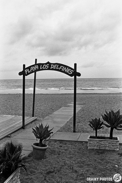 A sign reading "Playa Los Delfines" stands at the entrance of a sandy beach, with a path leading to the ocean. The scene is captured in black and white, highlighting the cloudy sky and coastal atmosphere.