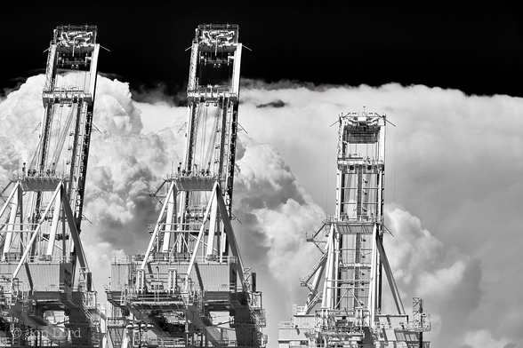 This is a high contrast black and white photo in landscape format of three raised gantry cranes in the foreground with large and developing storm clouds in the background. Long Beach, California (2015).

In the foreground are the three brilliant white giant gantry cranes (large cranes on the dockside used to load and unload shipping containers on and off container ships). One crane is in the lower left corner, its raised boom nearly reaching the top of the photo. The next crane is slightly to the left of centre, again, it's boom nearly touching the top of the image. The third crane is slightly to the right of centre and is either smaller or further away from the camera, the boom only rising to a little over the half way up level in the photo. Each crane sits on four legs extending upwards ten or more metres before there is a hinged boom that can be raised to allow a container ship to dock or lowered parallel to the ground enabling the shipping containers to be raised from the dockside, moved sideways over the ship and lowered onto or below the deck. In this photo the booms are raised upwards at an angle of about sixty degrees by means of cables fixed to the end of the booms, then to a stout 'A' frame like structure on top of the crane and then down to winches located in a large light grey corrugated enclosure stretching across under the 'A' frame. In the distance, perhaps thirty km away is a line of large developing cumulonimbus clouds. Larger on the left side and sloping downwards towards the right. The clouds are mostly a bright white with only a few grey areas adding to the contrast in the image. The clouds extend upwards from the base of the photo to just below the top. The cloud tops are, at a guess, around seven to ten thousand metres above the ground. The larger cloud on the left has the look of a giant cauliflower. Above is a clear and cloudless sky rendered jet black. 

The storms are viewed from the Port Of Long Beach looking east towards the San Gabriel mountains, which the storms are building up against. 