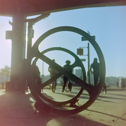 Round bike racks with horizontal beam in the middle back lit by the sun. There're a few cyclists in the background.