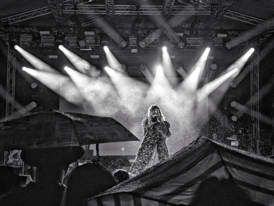 Black-and-white photograph of Mieze Katz, lead singer of German band MIA, performing at the centre of a rain-soaked stage. Dramatic beams of stage lighting cut through the darkness, illuminating her glittering costume as water droplets spray around her. In the foreground, silhouetted audience members hold umbrellas aloft. The stage features a visible metal truss structure with professional lighting fixtures, creating a striking contrast between the brightly lit performer and the shadowy, umbrella-filled crowd.