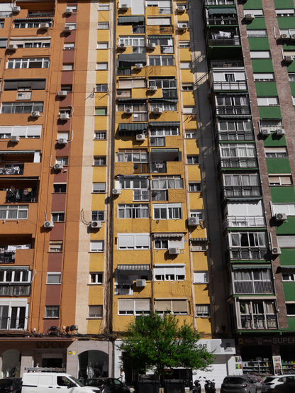 A daytime, front-facing view of a tall, apartment building facade. The building sections are painted in vibrant blocks of orange, yellow, and green.