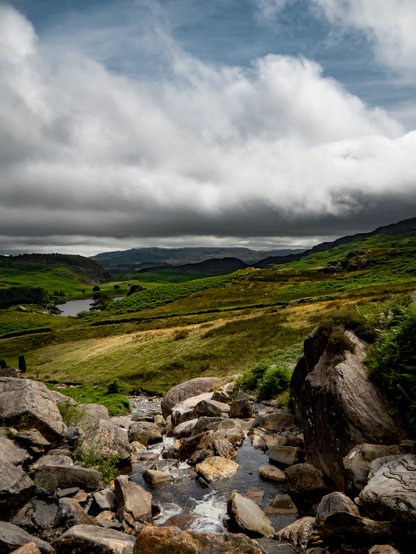 A landscape of a rocky stream leading to a Welsh valley on an overcast day. In the foreground, clear water rushes over large rocks and boulders towards a lush mountain valley with rolling hills in shades of green and yellow. A mountain lake can be seen in the distance while further on the mountains of Eryri (Snowdonia) rise to meet a dramatic sky with moody, low hanging clouds.