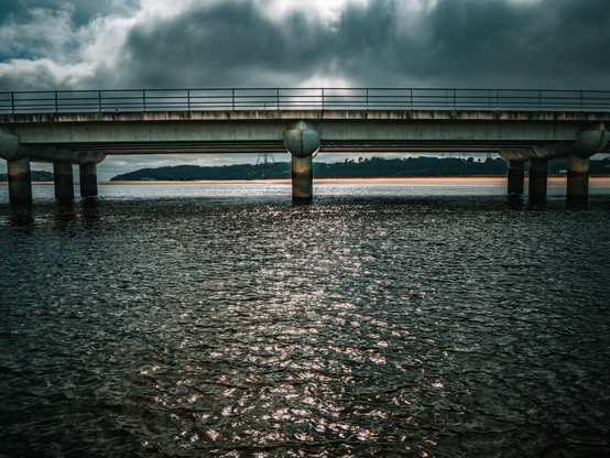 A landscape photo of a weathered concrete bridge with a metal railing crossing over a wide body of rippling water. Over the top of the bridge the sun is breaking through dark, moody clouds, casting a bright reflection down the centre of the water's surface. In the distance, a sandy shore and treeline are visible beneath the bridge structure.