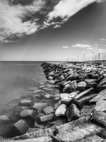 A rock wall next to a harbor is seen along the shore of Lake Huron. The large rocks that make up the wall are seen in detail as the wall juts out far into the large lake. The masts of sail boats can be seen to the right and large white clouds fill the sky. This is a black and white image.